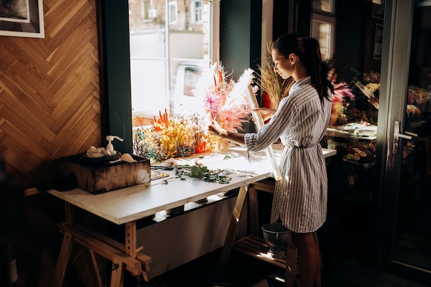 Un fleuriste vêtu d'une robe rayée se tient près de la table avec des fleurs séchées colorées et tient une enveloppe d'emballage transparente dans le fleuriste.