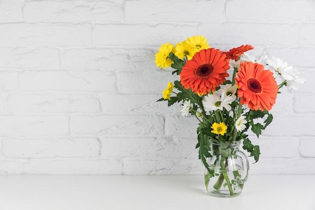 Fleurs de camomille et de gerbera dans le pichet en verre sur le bureau contre le mur de briques blanches