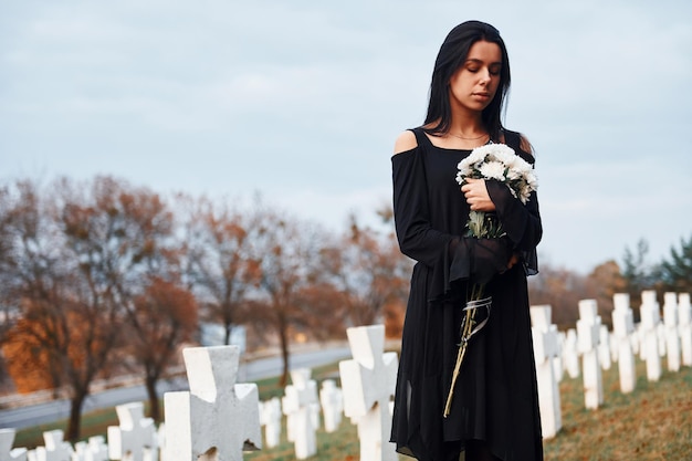 Avec des fleurs dans les mains Jeune femme en vêtements noirs visitant le cimetière avec de nombreuses croix blanches Conception des funérailles et de la mort