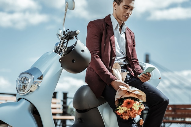 Avec des fleurs. Faible angle de gars agréable confiant transportant un bouquet de fleurs et s'appuyant sur une moto