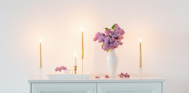 Fleurs lilas dans un vase blanc et bougies dorées sur fond de mur blanc