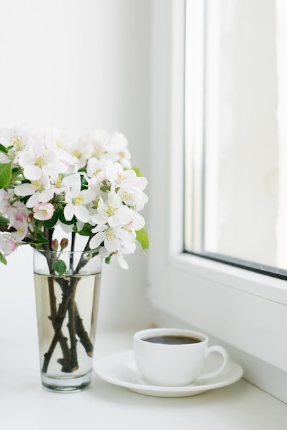 Fleurs de printemps dans un vase et une tasse de thé ou de café dans une tasse en porcelaine blanche sur le rebord de la fenêtre