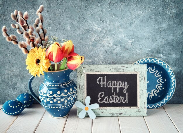 Fleurs de printemps en pichet en céramique bleue avec assiette assortie et oeufs de Pâques et un tableau noir.