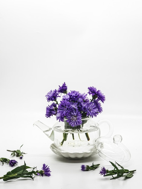 Fleurs violettes dans une théière en verre avec des feuilles sur la table.