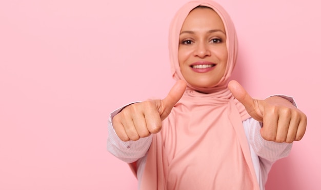 Flou artistique sur les mains d'un sourire flou avec un sourire à pleines dents Femme arabe musulmane en hijab rose, montrant les pouces vers la caméra, isolée sur fond coloré avec un espace pour le texte