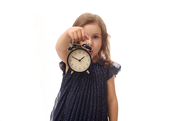 Flou artistique sur un réveil noir dans la main d'une jolie petite fille surprise en robe de soirée, posant sur fond blanc avec espace de copie. Concept de vendredi noir