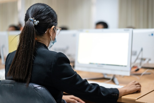 Flou et mise au point sélective des apprenants universitaires adultes portant un masque facial tout en se concentrant sur l'examen en ligne dans la salle informatique. Étudiants sérieux travaillant sur ordinateur à l'université