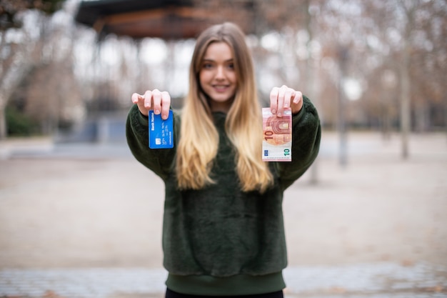 Floue jeune femme démontrant carte en plastique et de l'argent à la caméra tout en se tenant dans le parc