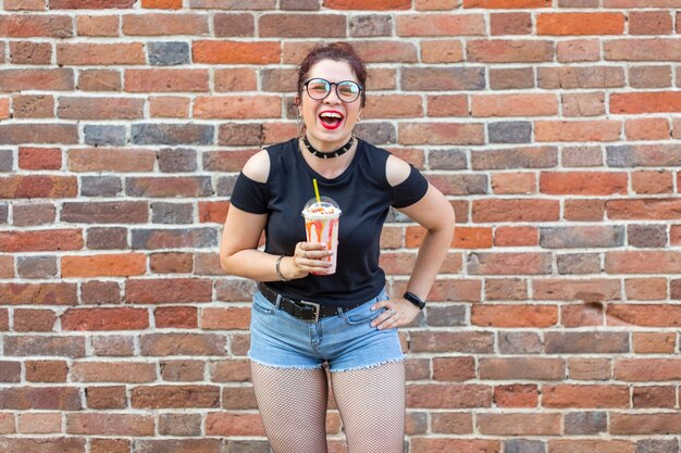 Floue jeune fille positive tient à Ruach un milkshake avec une paille contre un mur de briques. Concept de dessert d'été et week-end