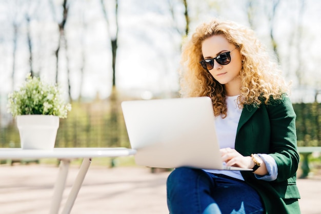 Fluffyhaired girl in sunglasses jeans veste verte livre de lecture messagerie en ligne avec ordinateur portable à l'extérieur engageant attrayant