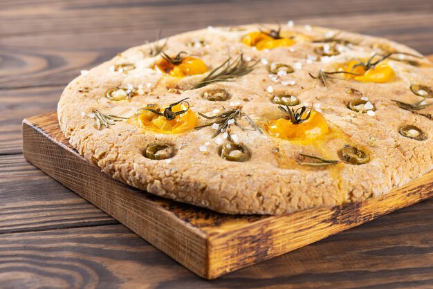 Focaccia italienne aux olives, tomates, romarin et épices sur une planche de bois.