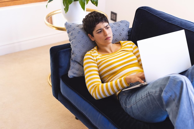 Focalisée femme biraciale allongée sur le canapé en utilisant un ordinateur portable à la maison espace de copie