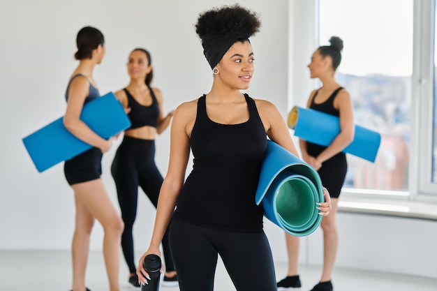Photo focus sur une femme afro-américaine heureuse debout avec un tapis de fitness et une bouteille de sport dans un studio de yoga