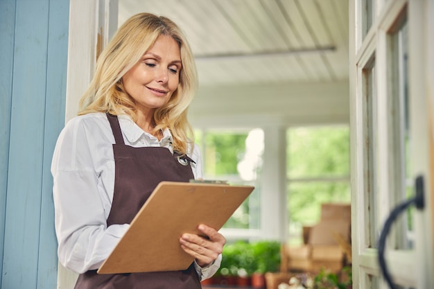 Focused heureux mature blonde femme caucasienne prenant des notes sur le presse-papiers dans sa main