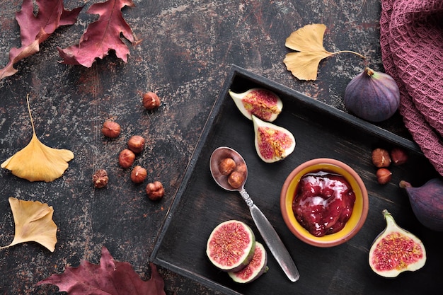 Fond d'automne avec des noix de cuillère à confiture de figues et des figues fraîches coupées en deux sur un plateau en bois noir Fond brun foncé avec des feuilles d'automne sèches