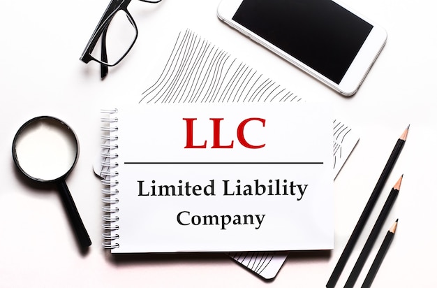 Sur un fond blanc des lunettes, une loupe, des crayons, un smartphone et un ordinateur portable avec le texte LLC Limited Liability Company