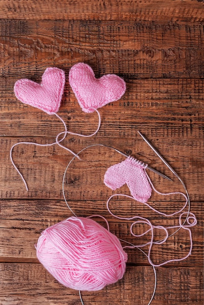 Sur un fond en bois, des coeurs roses tricotés, des outils à tricoter et du fil