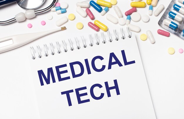 Sur un fond clair, des pilules multicolores, un stéthoscope, un thermomètre électronique et un cahier avec le texte MEDICAL TECH. Notion médicale.