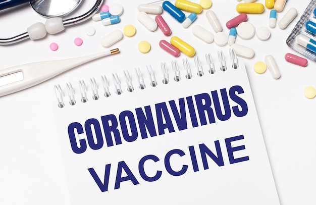 Sur un fond clair, des pilules multicolores, un stéthoscope, un thermomètre électronique et un cahier avec le texte VACCIN CORONAVIRUS. Notion médicale.