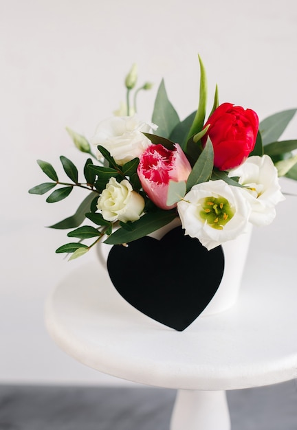 Fond de coeur de tableau noir. Bouquet de printemps dans un vase blanc sur un support blanc en bois. Roses, tulipes et lisianthus.