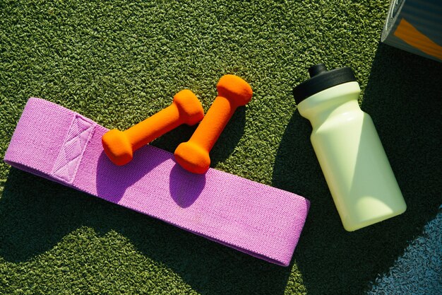 Sur le fond du gazon artificiel, il y a un tapis, une bouteille, une bande élastique de fitness et un tapis. Équipement sportif. Le concept d'un mode de vie sain, du sport et de l'alimentation. Une belle journée ensoleillée.