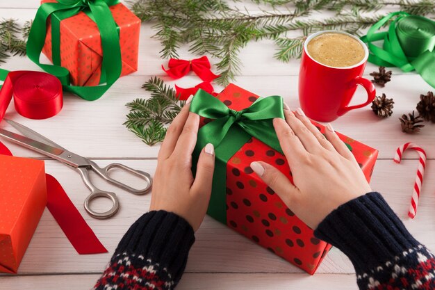 Fond d'emballage cadeau. Les mains des femmes emballent le cadeau de Noël dans du papier rouge, décorent avec un ruban de satin sur une table en bois rustique en désordre blanc. Concept de vacances d'hiver.