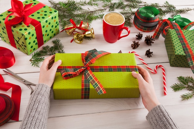 Fond d'emballage cadeau. Les mains des femmes emballent le cadeau de Noël en papier vert, décorent avec un ruban de satin sur une table en bois rustique en désordre blanc. Concept de vacances d'hiver.
