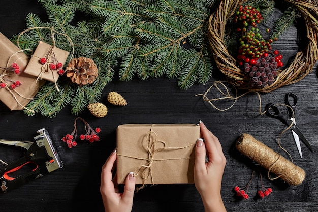 Fond de Noël. Vue de dessus des mains féminines envelopper le cadeau du Nouvel An. Cadeaux et parchemins emballés, branches d'épinette et outils sur table en bois. Lieu de travail pour la préparation de décorations faites à la main.