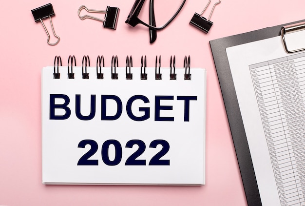 Photo sur fond rose, des rapports, des trombones noirs, des lunettes et un cahier blanc avec le texte budget 2022.