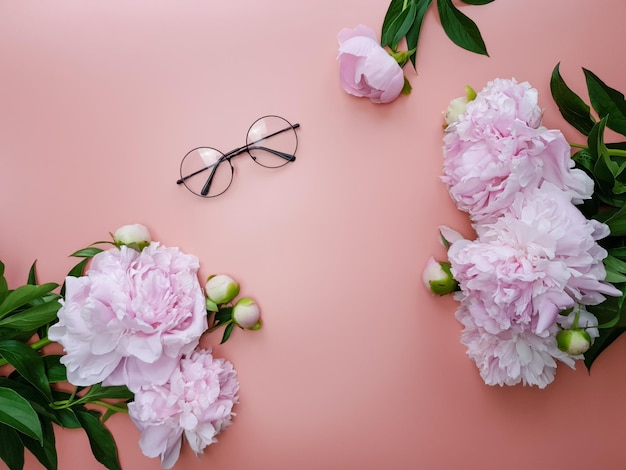 Sur un fond rose se trouve une paire de lunettes et des pivoines roses