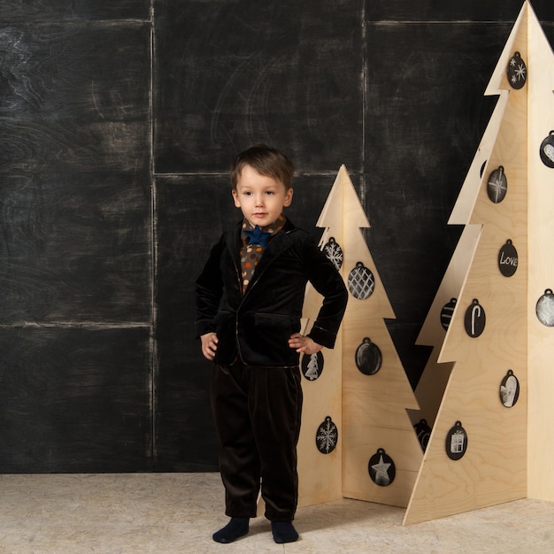 Sur un fond sombre petit garçon posant dans un costume élégant près d'un arbre de Noël en bois