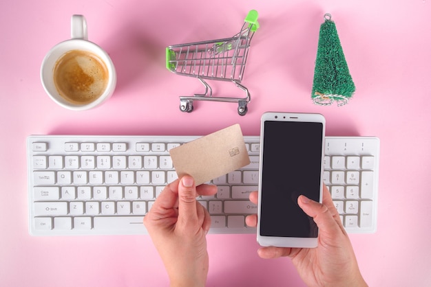 Fond de vente de Noël vendredi noir avec des sacs en papier vert rouge, panier, carte bancaire, clavier d'ordinateur, smartphone dans les mains d'une fille vue de dessus flatlay