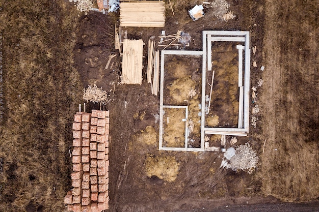 Fondation en béton pour le sous-sol de la future maison, des piles de briques et des rondins de bois de construction pour la construction le jour d'été ensoleillé, vue aérienne.