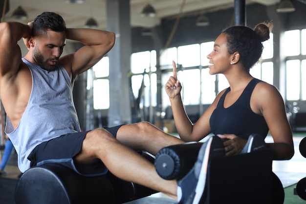 Formateur aidant un jeune homme à faire des exercices abdominaux dans une salle de sport.