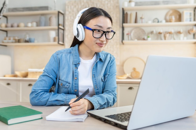 Formation en ligne jeune belle fille asiatique assise à la table à la maison dans un casque blanc et avec