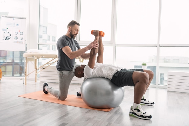 Formation en rééducation. Bel homme agréable allongé sur le medball tout en soulevant des haltères