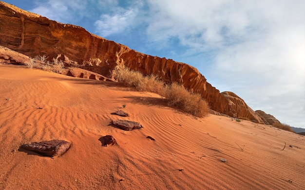 Formations rocheuses dans le désert de Wadi Rum, le soleil brille sur le sable rouge, les rochers, quelques buissons secs, le ciel bleu au-dessus