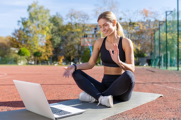 Formatrice en ligne assise sur un tapis de sport dans un stade pendant la journée en utilisant un ordinateur portable pour la vidéo