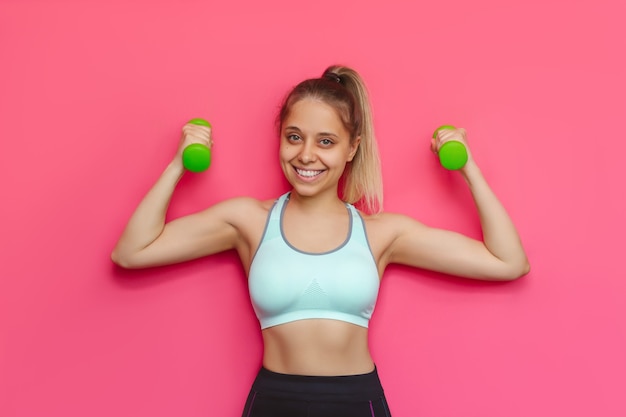 Forte belle jeune femme blonde caucasienne avec des sourires d'haltères verts isolés sur un mur rose vif et coloré