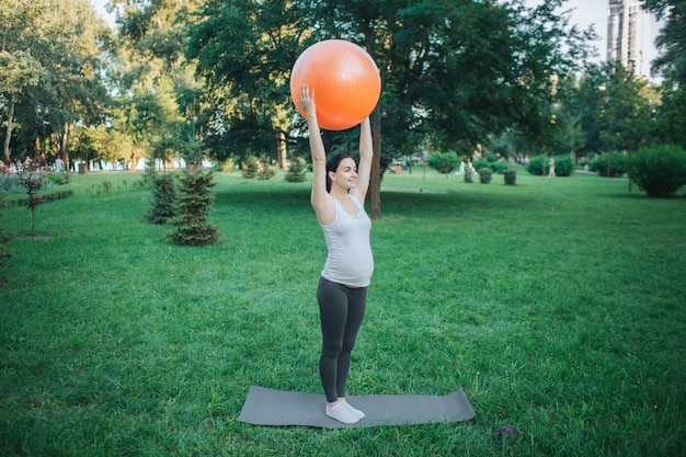 Forte femme enceinte oyung exerçant sur yoga mate dans le parc. Elle tient un gros ballon de fitness orange dans les mains au-dessus de la tête.