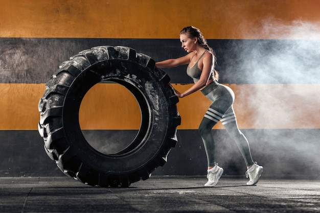 Forte jeune femme athlétique soulevant un gros pneu dans le fond sombre de la salle de gym.