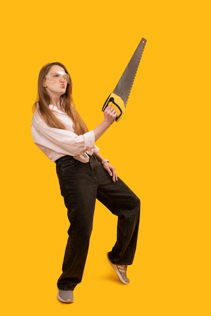 Forte jeune femme portant des lunettes et tenant une scie à main posant pour la photo Isolé sur fond jaune Cadre vertical Copier l'espace