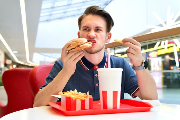 Un fou affamé mord en mangeant un hamburger de malbouffe rapide, des frites et des boissons gazeuses. Faim