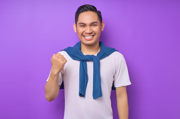 Fou de joie et excité jeune homme asiatique des années 20 portant un t-shirt blanc faisant un geste gagnant célébrer les poings serrés dire oui isolé sur fond violet concept de style de vie des gens