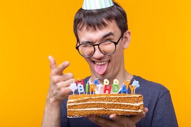 Fou joyeux jeune homme avec des lunettes tenant une bougie allumée dans ses mains et un gâteau de félicitations sur un mur jaune. Concept de célébration anniversaire et anniversaire.