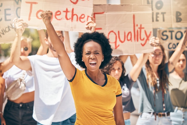 Une foule de manifestants et une femme noire dans la rue poing dans l'air marchant pour l'égalité, les droits de l'homme et la liberté Manifestant pour la diversité et une femme criant pour la justice dans la ville avec des panneaux en carton