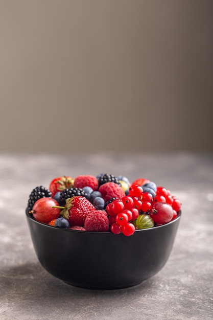 Fraise fraîche, mûre, groseille à maquereau, framboise et myrtille dans un bol noir avec sur gris.