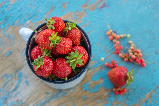 Photo des fraises juteuses fraîches dans une tasse en émail sur une vieille table rurale en bois à côté se trouvent des baies de cassis