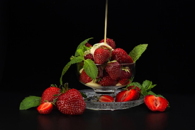 Fraises mûres dans un vase en verre avec des feuilles vertes à la crème avec des fraises coupées posées sur du verre dépoli sur fond noir