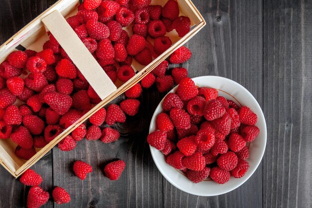 Photo des framboises fraîches mûres dans un panier et dans un bol sur un fond en bois noir